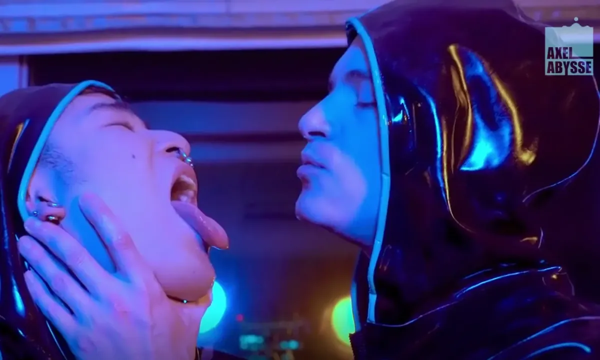 Axel Abysse still: two performers in glossy latex hoods under neon light